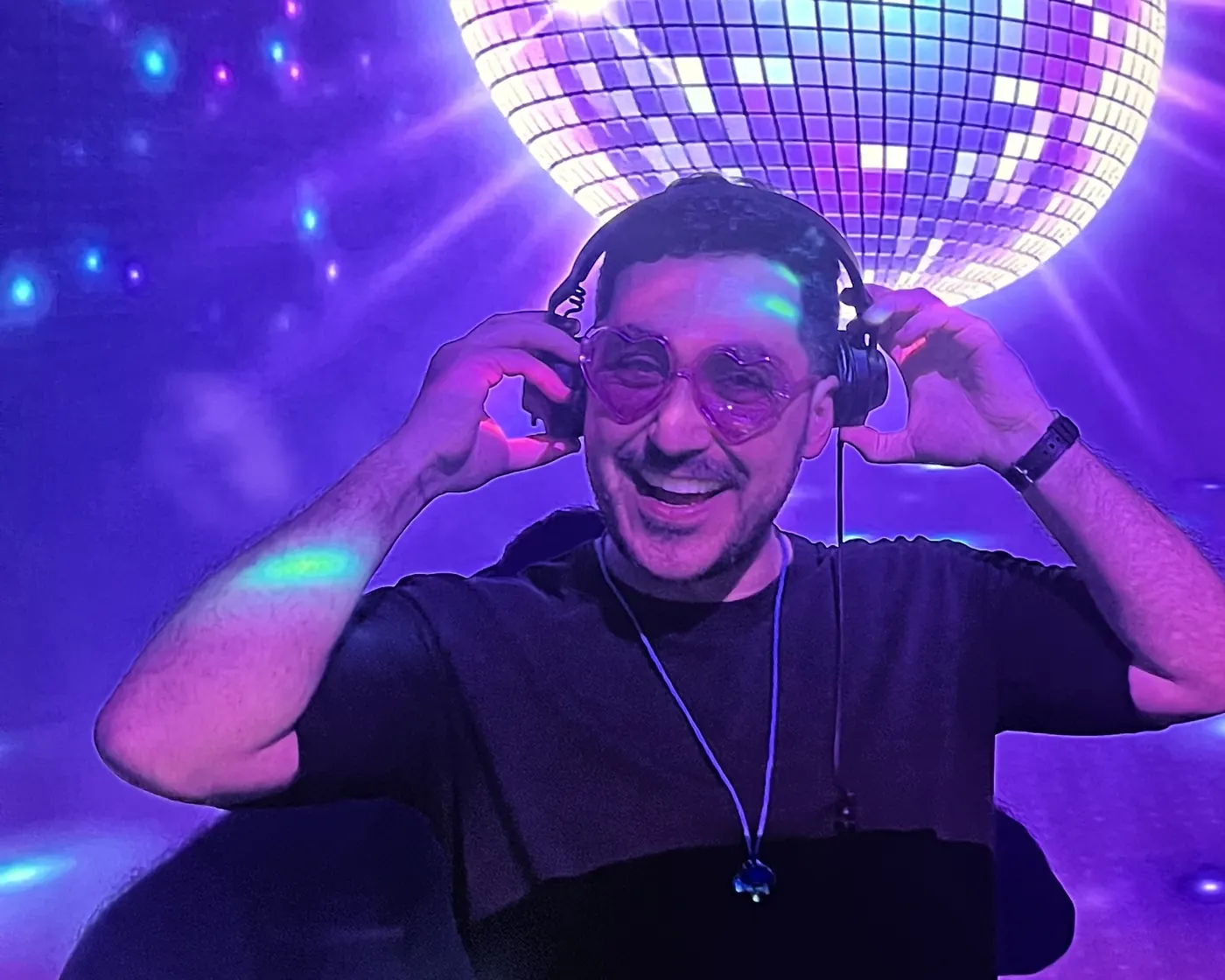 Avand behind the decks, headphones in both hands, a mirror ball lit up above him